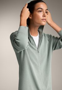 Zelena kapuco z zadrgo iz teksturirane tkanine z rebrastimi manšeti in visokim ovratnikom, z sprednjo zadrgo in oprijeto obliko.