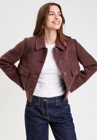 Braune strukturiertes kurzes Jackett mit Kragen, großen Knöpfen und vordertasche. Getragen über einem weißen Hemd und blauen Jeans.