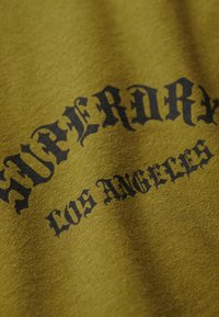 Oliwkowy bawełniany t-shirt z wyraźnym czarnym napisem "SUPERDRY LOS ANGELES" w ozdobnej czcionce na przodzie.