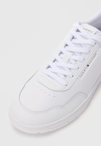 Zapatilla blanca con parte superior sintética lisa, puntera redonda, lengüeta acolchada y cordones planos. Presenta perforaciones y un logo lateral.