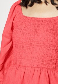 Blusa coral com decote quadrado, textura franzida no corpete e mangas longas com bainha franzida. Feita de um tecido leve.
