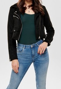Zwarte suède cropped jas met rits- en zakdetails, gecombineerd met aansluitende blauwe denim jeans en een donkergroene top.