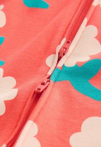 Közelkép korallszínű anyagról, fehér és tengerzöld virágmintával, valamint két korallszínű cipzárhúzóval, amelyek egy részben zárt cipzáron vannak.