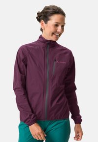 Personne souriante portant une veste zippée bordeaux avec une poche poitrine et un pantalon bleu-vert, se tenant devant un fond clair.