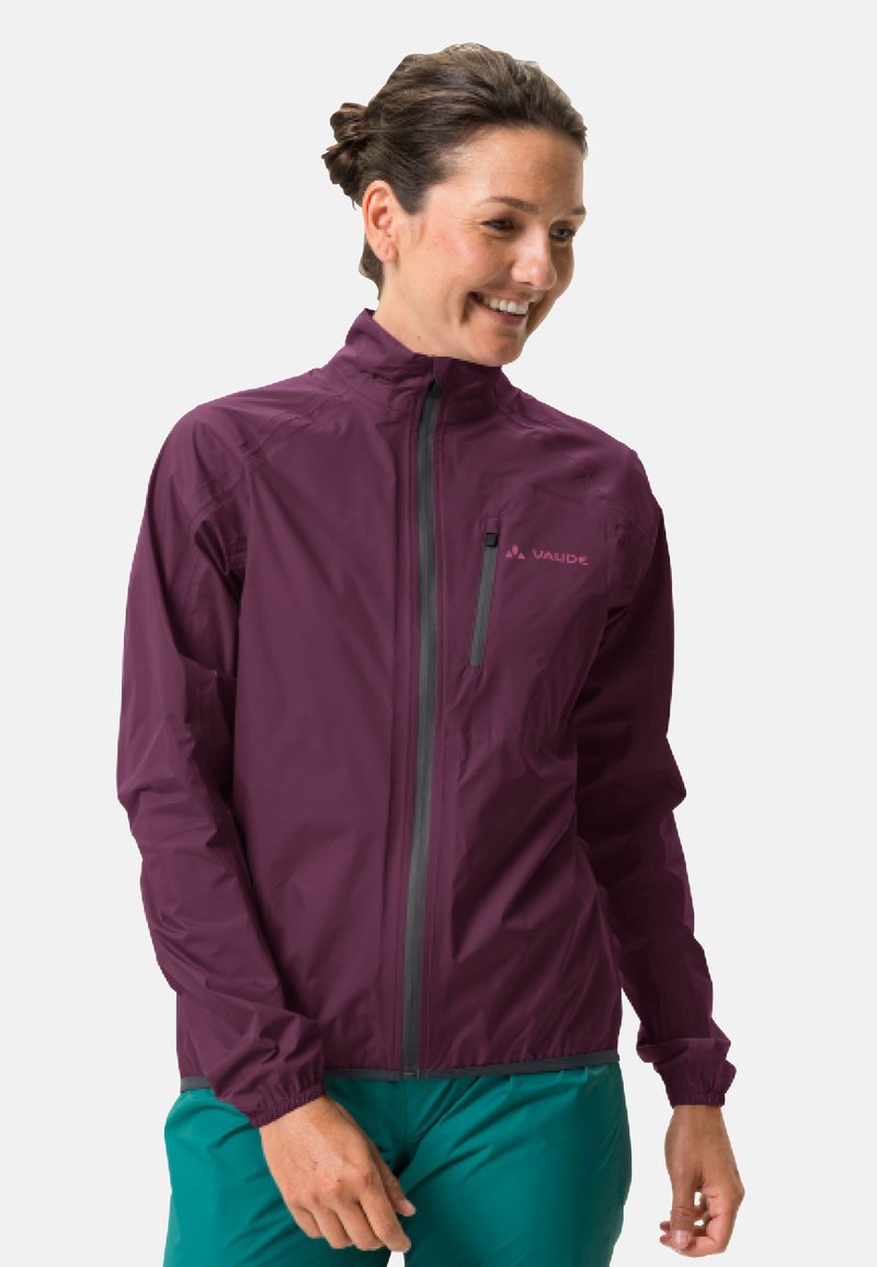 Personne souriante portant une veste zippée bordeaux avec une poche poitrine et un pantalon bleu-vert, se tenant devant un fond clair.