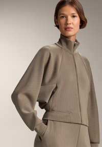 Svetlo béžová zipsová bunda s vysokým golierom, rebrovanou textúrou a širokými, voľnými rukávmi. Obsahuje vertikálne prúžky a priliehavý pás.