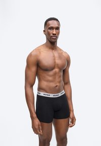 Άνδρας με γυμνό στήθος και μυώδη κορμί, φορώντας μαύρα boxer briefs Hugo, σε απλό φωτεινό φόντο, όρθιος και κοιτάζοντας μπροστά.