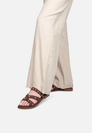 Persoon draagt beige broek met wijde pijpen en bruine platte sandalen met studs, staand tegen een effen achtergrond.
