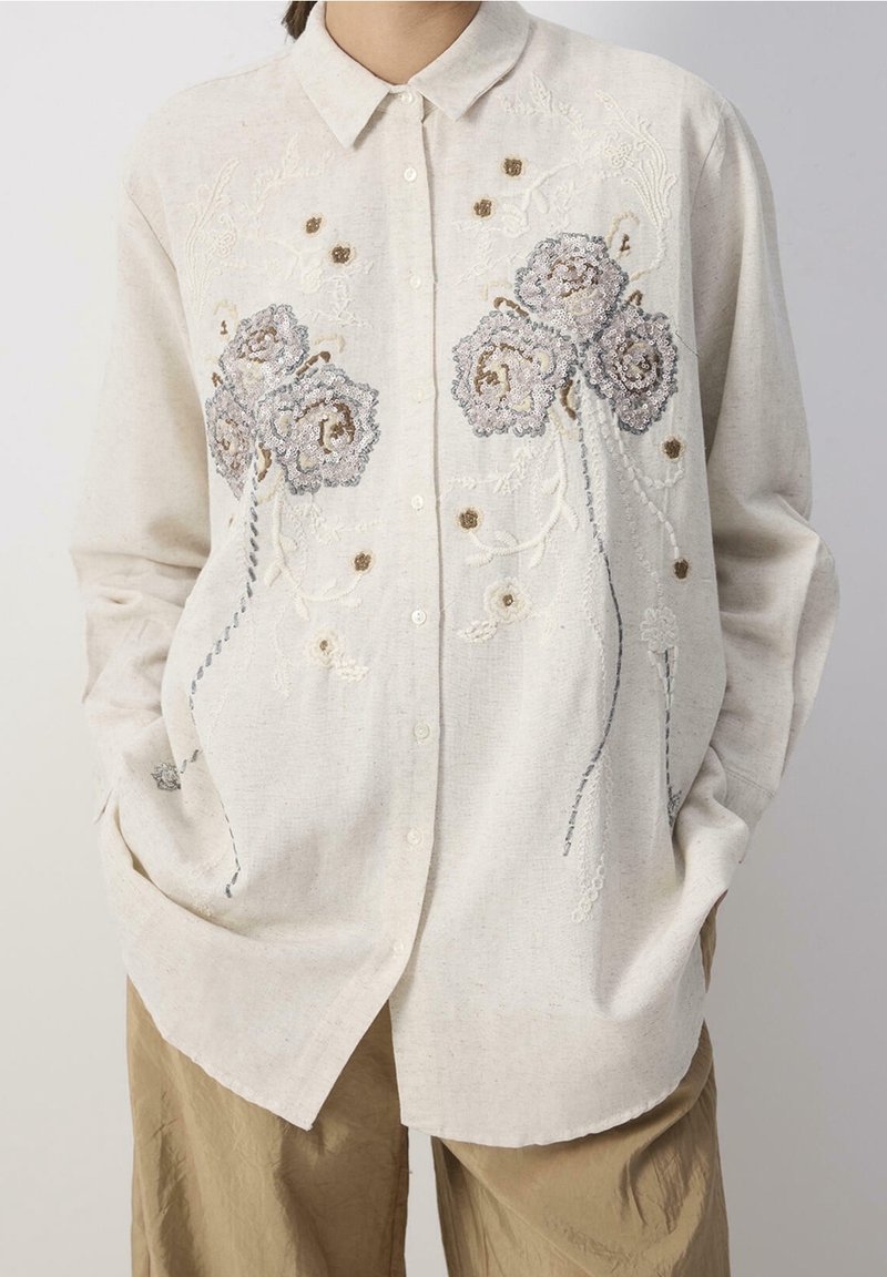 Femme portant une chemise beige à boutons avec des motifs floraux brodés et pailletés, associée à un pantalon marron clair, mains dans les poches.