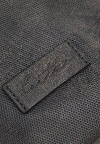 Pelle nera strutturata con una toppa rettangolare liscia con scritte a rilievo. Cuciture visibili intorno alla toppa.