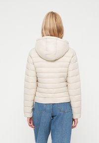 Lichtbeige gewatteerde jas met capuchon, voorzien van horizontale gestikte secties, een gladde textuur en een cropped ontwerp. Gecombineerd met blauwe denim.