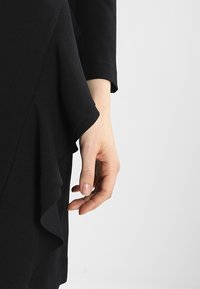 Abito nero con orlo a volant. Una mano è delicatamente poggiata sul tessuto, che appare morbido e leggermente testurizzato. Maniche lunghe visibili.