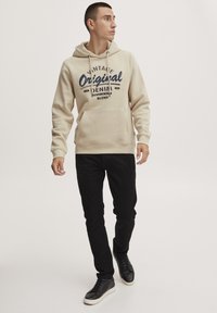 Sudadera crema con un bolsillo frontal y estampado gráfico en color navy, combinada con pantalones negros y zapatillas negras. Tela suave y diseño informal.