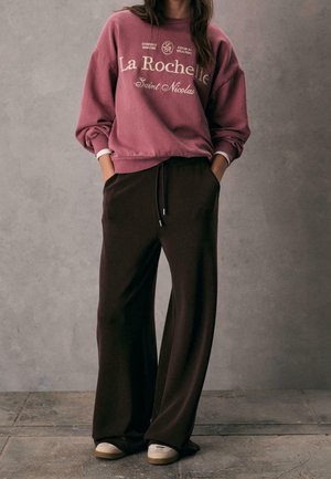 Personne portant un sweat mauve avec le texte « La Rochelle Saint Nicolas », un pantalon large foncé et des baskets blanches, se tenant contre un mur gris.