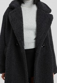 Zwarte textuur oversized jas met sjaalkraag, voorzien van grote zwarte knopen en een zachte, pluizige stof, gedragen over een lichtgrijze coltrui.