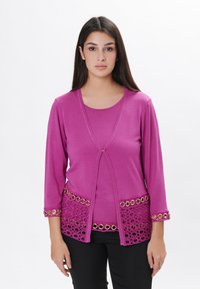 Fuchsia Strickjacke mit Dreiviertelärmeln, verziert mit Spitzenbesatz und goldenen Ösenakzenten am Saum und den Bündchen, kombiniert mit einem passenden Oberteil.