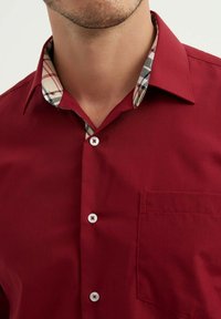 Chemise rouge à boutons, avec un col à motif à carreaux, présentant des boutons blancs, une poche au côté gauche de la poitrine, et une texture de tissu lisse.