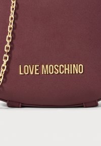 Burdona rokassoma ar zelta toņa "LOVE MOSCHINO" logo. Iezīmēts ar ķēdes siksniņu, gludu tekstūru, noapaļotu formu un minimālistisku dizainu.