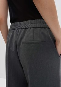 Persona che indossa pantaloni grigio scuro con vita elasticizzata e tasca posteriore, abbinati a una camicia nera infilata dentro.