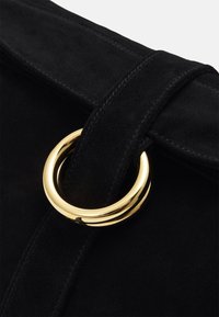 Sac à main en suède noir avec un accent de quincaillerie circulaire dorée, présentant un rabat structuré et des détails de couture.