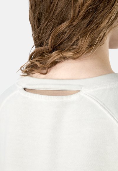 Persona con cabello castaño claro ondulado que lleva una blusa blanca con un corte horizontal estrecho en la parte posterior del escote.