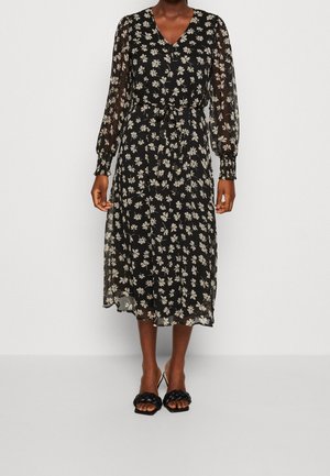 Sort midi-kjole med beige blomsterprint, gennemskinnelige lange ærmer, V-hals, indsnævret talje bundet med bælte, båret med sorte hælede sandaler.