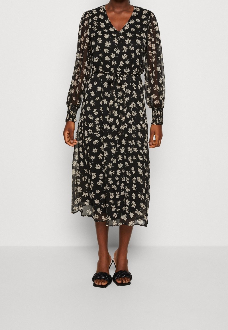 Robe midi noire avec imprimé floral beige, manches longues transparentes, col en V, taille cintrée avec ceinture, portée avec des sandales à talons noires.