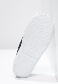 Zapatilla de lona azul marino con una suela de goma blanca texturizada, que presenta una puntera redonda y plana, y un logo grabado en el costado de la suela.