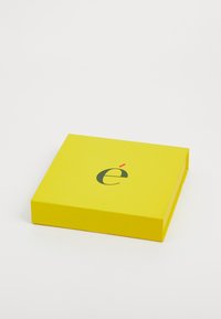 Scatola di cartone gialla con superficie liscia, caratterizzata da una lettera "é" nera stilizzata e da un piccolo accento rosso. Forma quadrata, chiusa.