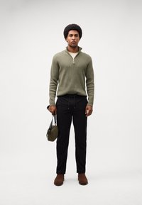 Olivengrønn zippered genser, lys skjorte under, svarte bukser, brune sko, holder en liten olivengrønn veske, stående foran en ensfarget bakgrunn.