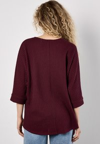 Vrouw met lang, krullend blond haar, gekleed in een losse, gestructureerde bordeauxrode trui en lichtblauwe jeans, met de rug naar de camera tegen een effen achtergrond.