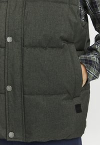 Dark green puffer vest made of textured fabric, featuring silver snap buttons, side pockets, and a small black brand label on the hem.