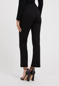 Kobieta w czarnych spodni wysokiej talii i czarnych sandałach na obcasie z paskami na kostkach, stojąca na tle białym.