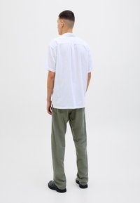 Hombre con pelo corto y oscuro, vistiendo una camisa blanca de manga corta holgada, pantalones verde oliva y zapatos negros, de pie de espaldas sobre un fondo liso.