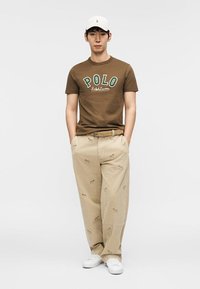 Uomo con cappellino bianco, t-shirt marrone "POLO Ralph Lauren", pantaloni beige con stampe di cani e sneakers bianche, in piedi con le mani nelle tasche.