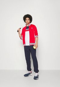 Red track jacket with white and navy stripes, white T-shirt, navy pants, holding a tennis ball and racket, finished with blue sneakers.