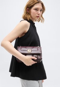 Γυναίκα με μαύρο τοπ χωρίς μανίκια που κρατά μια σκούρα καφέ δερμάτινη τσάντα με χρυσό κούμπωμα μπροστά σε απλό ανοιχτό φόντο.