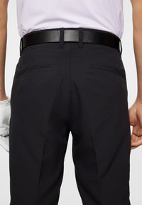 Svarta träningsbyxor med en slät yta, med två bakfickor och ett svart bälte. En vit handske kikar fram från vänster hand.