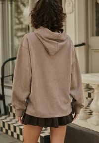 Brązowy welurowy hoodie z kapturem, w luźnym kroju, z prążkowanymi mankietami, w połączeniu z czarną plisowaną spódnicą, stojąc na szachownicowej podłodze.