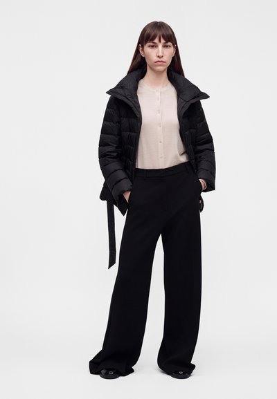 Femme debout les mains dans les poches, portant une chemise beige boutonnée, un pantalon noir à jambes larges et une veste noire matelassée avec le col relevé.