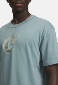 Heled sinine puuvillane T-särk lühikeste varrukatega, millel on suur allergia valgest ja mustast siidist tikitud logo, millel on tekstuuriga houndstooth mustri detail.