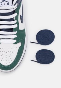 Gröna och vita högsnörda sneakers med överdel av l äder, perforerat tåparti och vita snören. Två marinblå snören ligger bredvid skon.