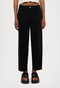 Pantalones de pierna ancha negros, hechos de algodón con un ajuste relajado, que cuentan con un cierre de botón frontal y bolsillos laterales, combinados con sandalias negras gruesas.