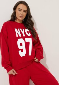 Sudadera roja de gran tamaño hecha de algodón con impresión blanca "NYC 97". Cuenta con puños y dobladillo acanalados, ajuste holgado, combinada con pantalones a juego.