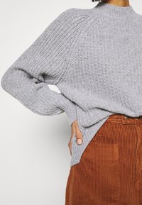 Světlě šedý pletený svetr s vysokým límcem a žebrovanou strukturou, kombinovaný s rezavě zbarvenou manšestrovou sukní s předními kapsami.