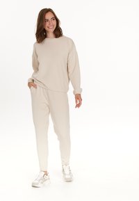 Beige matchande sweatshirt och joggers-set med avslappnad passform, elastiska ärmar och sidofickor, parat med vita sneakers.