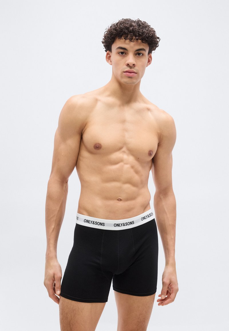 Muskulöser Mann trägt schwarze Boxer-Shorts mit weißem Bündchen, auf dem ONLY&SONS steht, und steht vor einem schlichten hellgrauen Hintergrund.