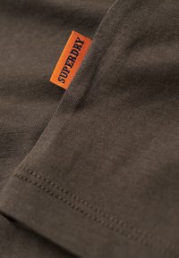 Camiseta de algodón marrón con textura acanalada. Presenta una etiqueta naranja que muestra "SUPERDRY". Detalles de costura visibles a lo largo del dobladillo.