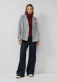 Grijze dubbelzijdige jas met gouden knopen, gedragen over een bordeauxrode coltrui. Gecombineerd met donkere wijde jeans en beige schoenen.
