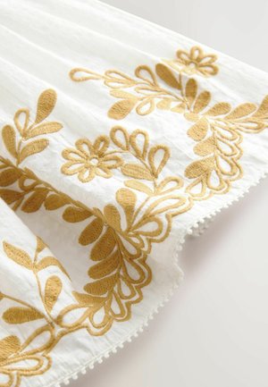Tissu blanc avec broderies florales dorées et motifs de feuilles, présentant une finition texturée et des bords scallopés décoratifs.